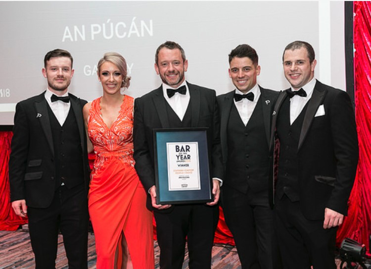 An Púcán Team Wins Bar of the Year Award – A group of five formally dressed individuals, including a woman in a red gown, celebrate on stage as An Púcán wins the "Bar of the Year" award. A large screen behind them displays the venue's name.
