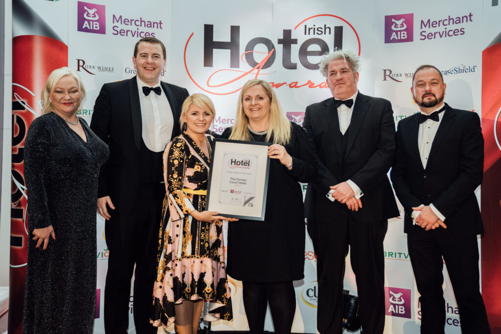 Group of Hotel Professionals Receiving an Award at the Irish Hotel Awards – A team of six elegantly dressed individuals, including men in tuxedos and women in formal attire, stand together holding an award at the Irish Hotel Awards event. The backdrop showcases the event sponsors and branding.
