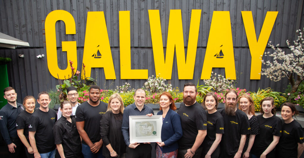 Restaurant Team Posing with an Award in Front of a "GALWAY" Sign – A group of hospitality staff, dressed in black t-shirts, stand together outside a venue featuring a bold yellow "GALWAY" sign, holding a framed award. The backdrop includes flowers and greenery, adding to the celebratory atmosphere.