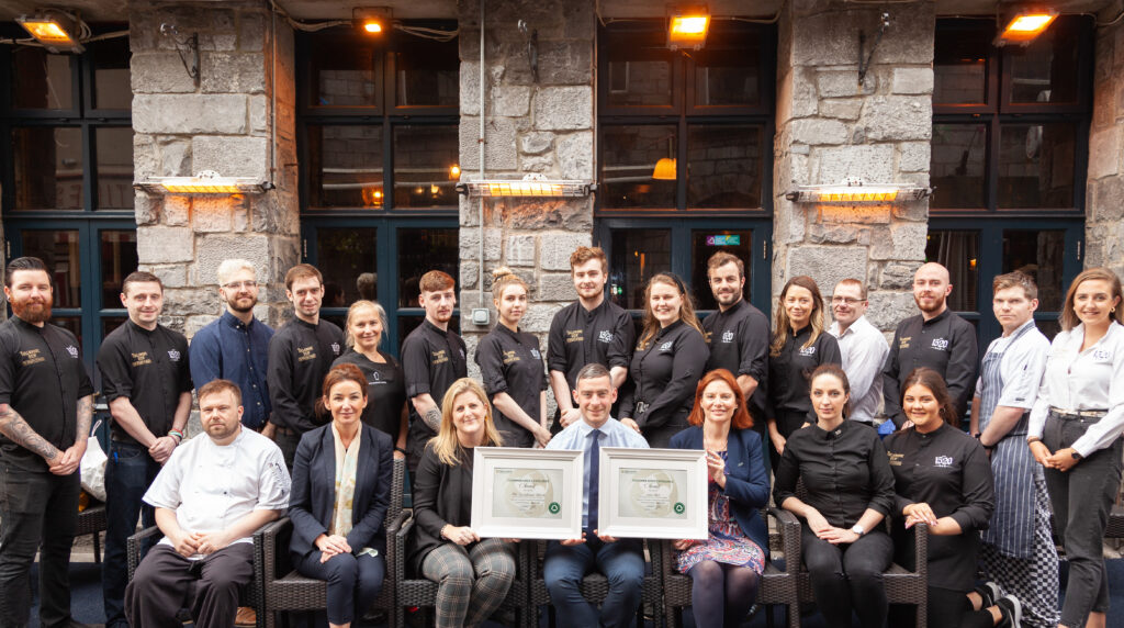 Restaurant Team Holding Awards Outside a Rustic Stone Building – A team of restaurant staff and management, dressed in a mix of formal and chef attire, pose in front of a rustic stone building, proudly holding framed awards. The warm lighting and classic architecture add to the celebratory ambiance.
