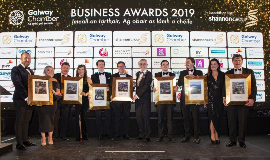 Winners at the Galway Chamber Business Awards 2019 – A group of award recipients, dressed in formal attire, pose on stage holding framed awards at the Galway Chamber Business Awards 2019. A backdrop features event sponsors and the chamber's branding.