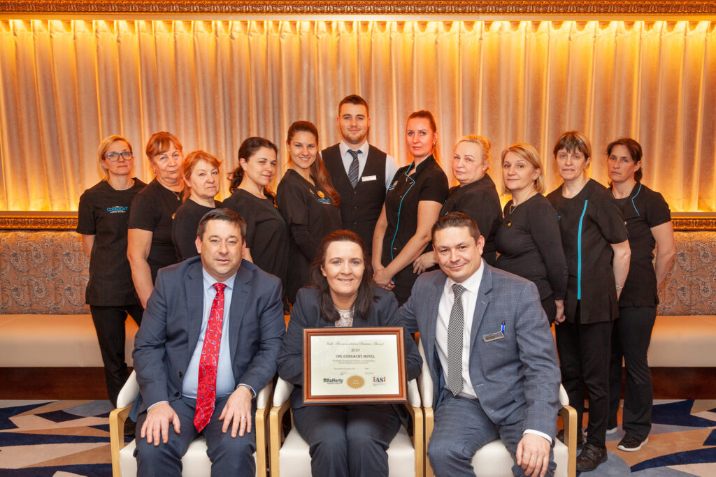 Hotel Staff and Management Holding an Award Indoors – A group of hotel employees, including management and housekeeping staff, pose inside an elegant lounge area. A woman in the center holds a framed award, highlighting the team's achievement.