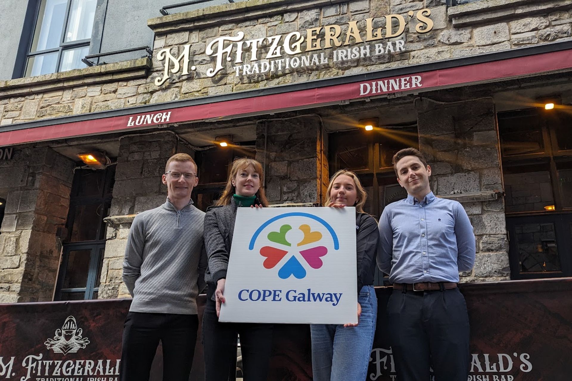 COPE Galway Partnership at M. Fitzgerald’s Bar – Four individuals stand in front of M. Fitzgerald’s Traditional Irish Bar, holding a COPE Galway sign, showing support for the organization’s community initiatives.