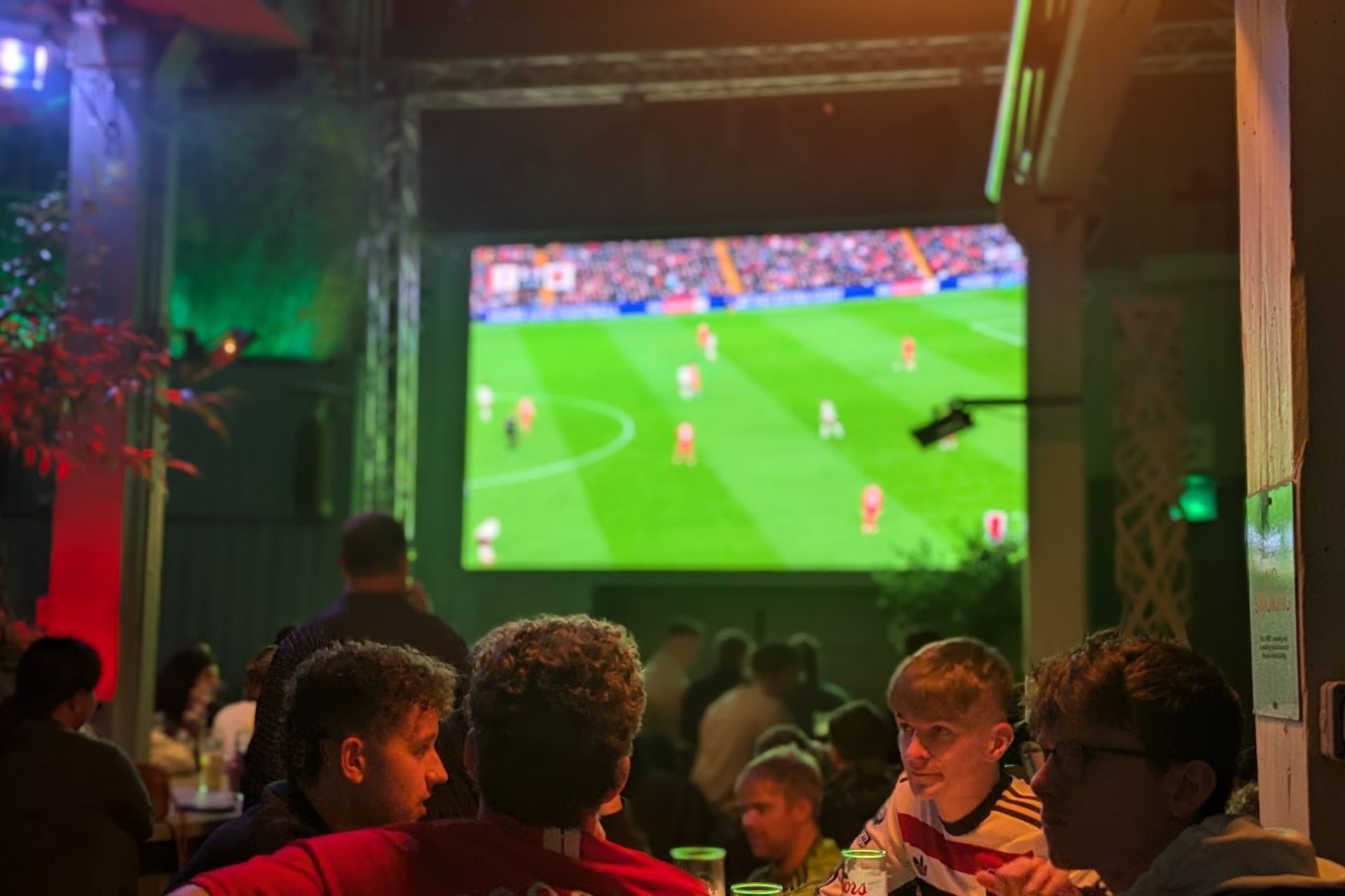 Sports fans watching a football match – A group of young men gathered at a lively venue watch a football game on a large screen, surrounded by vibrant lighting and an energetic atmosphere.