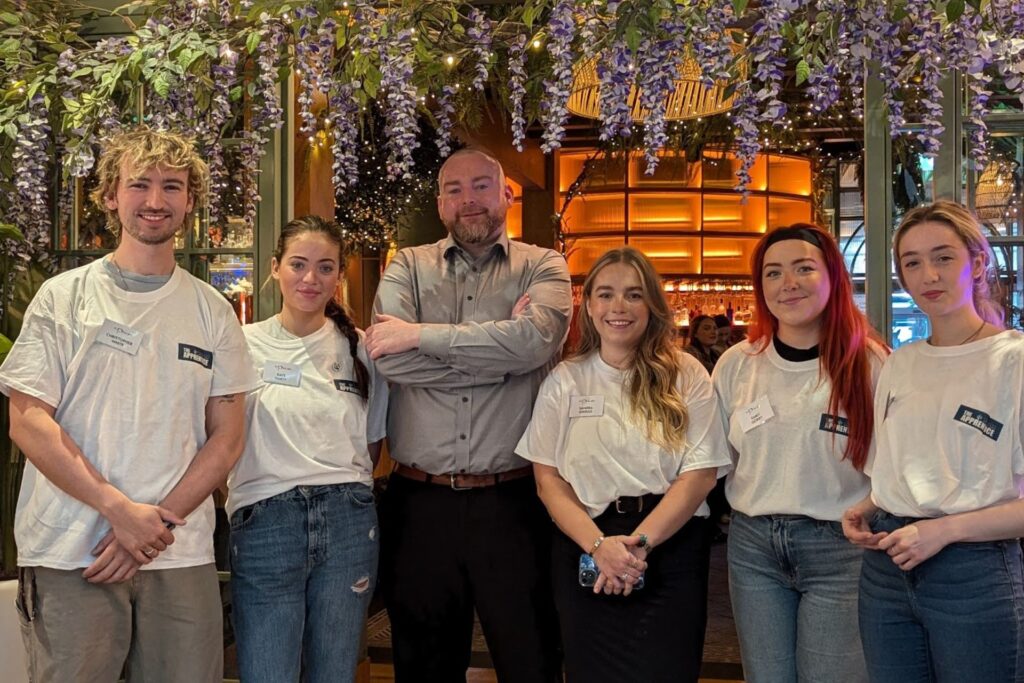 Group of staff members in a hospitality venue – A team of six individuals, five wearing white branded t-shirts and one in a grey shirt, pose together under hanging purple flowers in a stylish bar or restaurant with warm lighting.