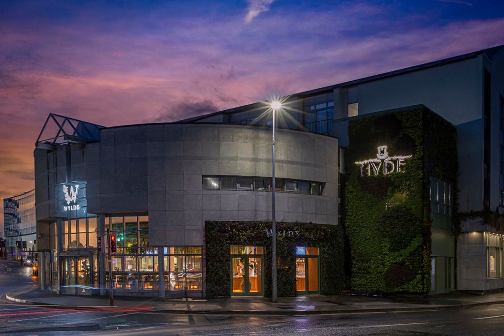 A stylish, contemporary building with illuminated signage for "HYDE" and "WYLDE," featuring a green wall and large glass windows, set against a twilight sky.