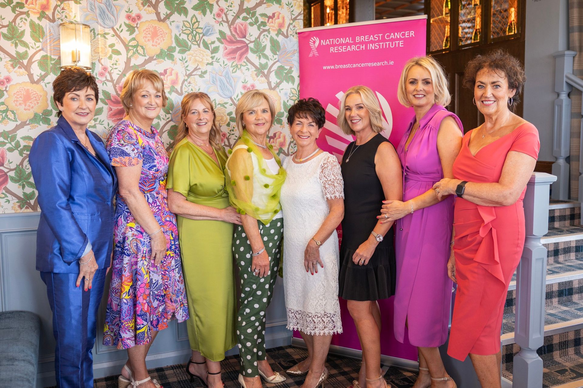 Glamorous Breast Cancer Research Event – Eight elegantly dressed women pose at a National Breast Cancer Research Institute event, standing in front of a floral wall and a pink banner promoting breast cancer research.