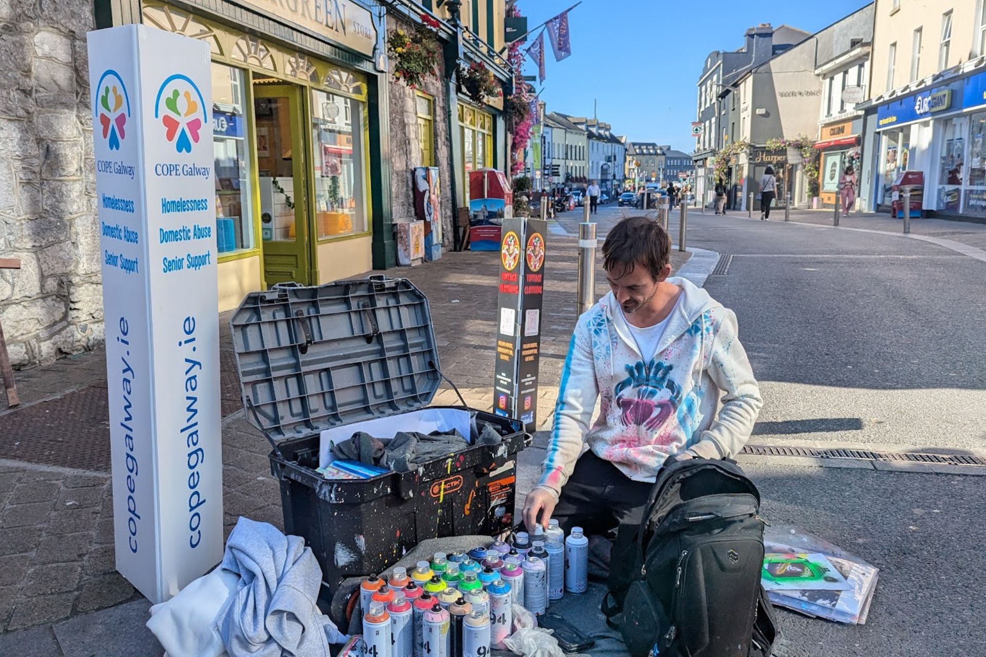 Street Artist Supporting COPE Galway – A street artist sits on a sidewalk with spray paint cans, preparing a mural, next to a COPE Galway sign raising awareness for homelessness, domestic abuse, and senior support.