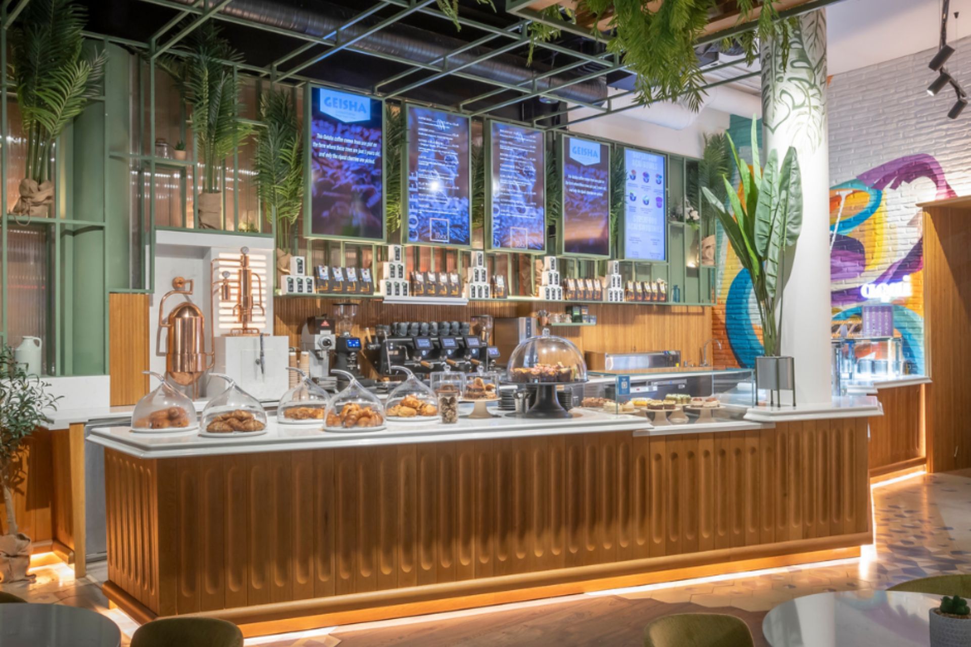 Modern Café Counter at WYLDE with Pastries and Coffee – A stylish café with a wooden counter, a variety of pastries on display, and digital menu boards, complemented by a vibrant, plant-filled interior.