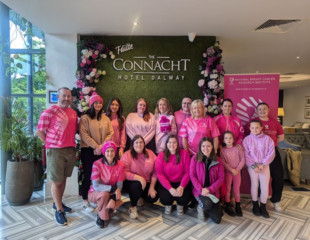 Pink-Themed Fundraiser at The Connacht Hotel Galway – A large group of women, men, and children dressed in shades of pink pose in front of The Connacht Hotel Galway's floral backdrop, supporting the National Breast Cancer Research Institute.