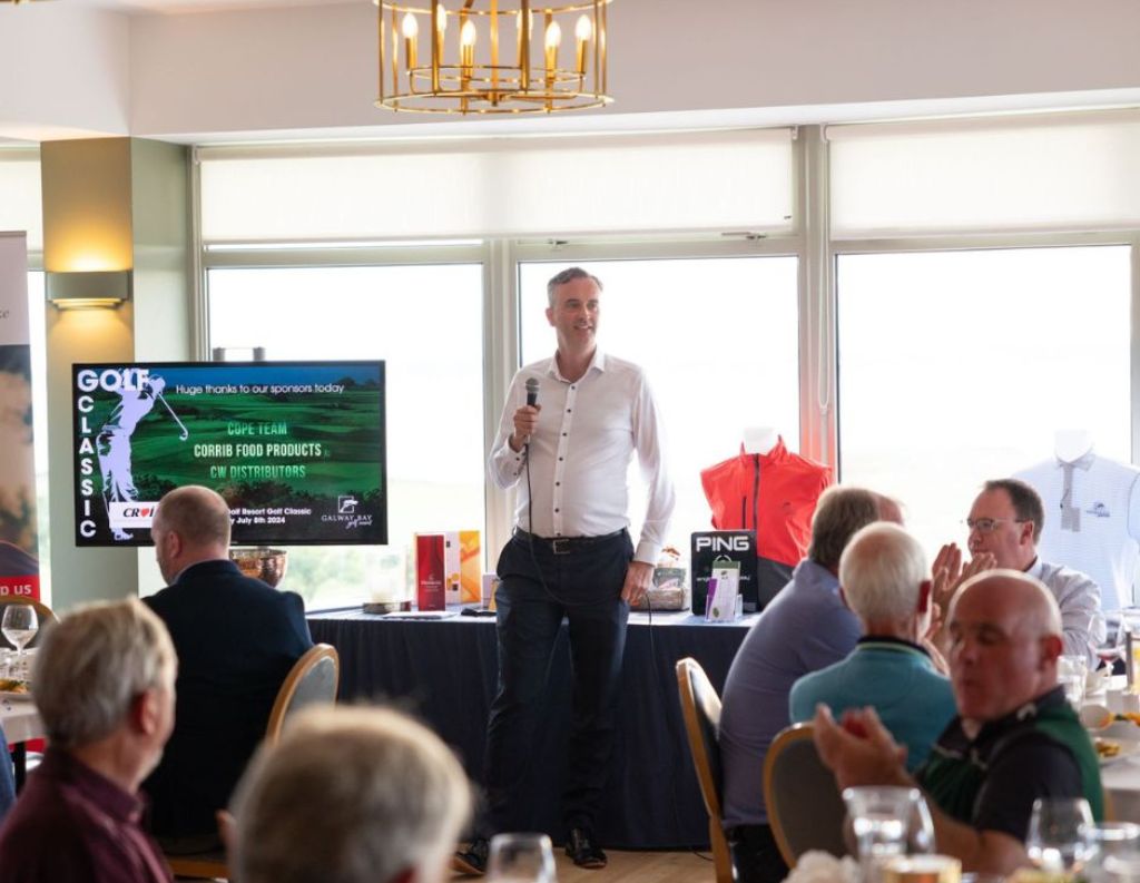 Golf Event Speaker Addressing Audience – A man in a white shirt speaking into a microphone at a golf event, addressing a room of seated attendees with banners and prizes in the background.