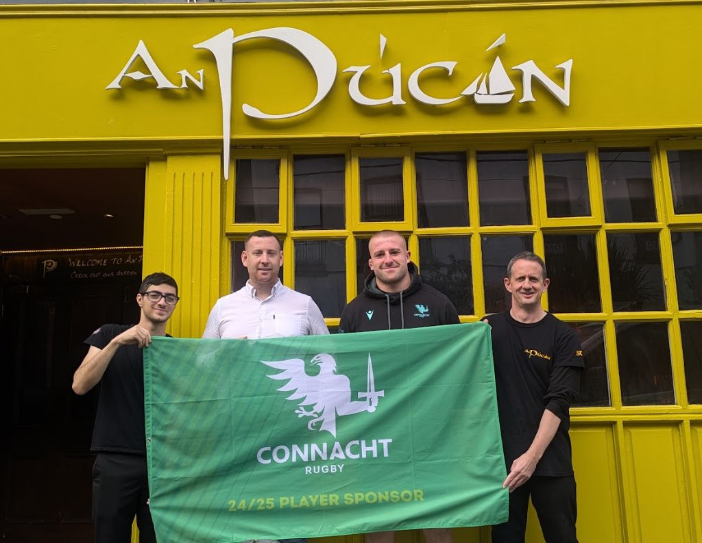 An Púcán Team Holding a Connacht Rugby Flag – Four men stand outside An Púcán, a vibrant yellow pub, holding a green Connacht Rugby flag. They are dressed in casual and team attire, showing support for the rugby team.