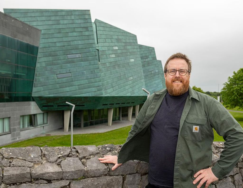 Man Posing in Front of a Modern Green Copper-Clad Building – A bearded man wearing glasses and a green jacket leans against a stone wall, smiling in front of a distinctive modern building with an angular copper-clad facade, set against a cloudy sky.