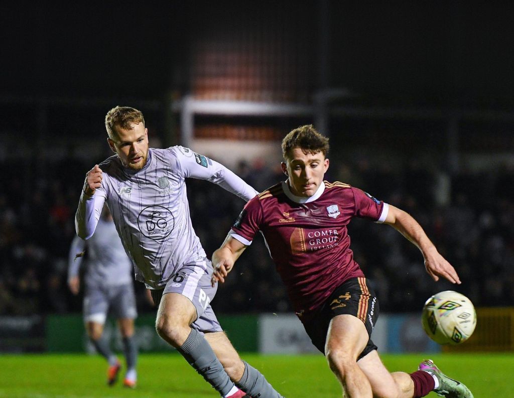 Two soccer players in action during a nighttime match. One player, wearing a maroon Galway United jersey with the Connacht Hospitality Group logo, is sprinting towards the ball with determination. The opposing player, dressed in a light gray kit, is closely following, trying to challenge for possession. Stadium lights illuminate the scene, highlighting the intensity of the game, with a blurred crowd in the background.