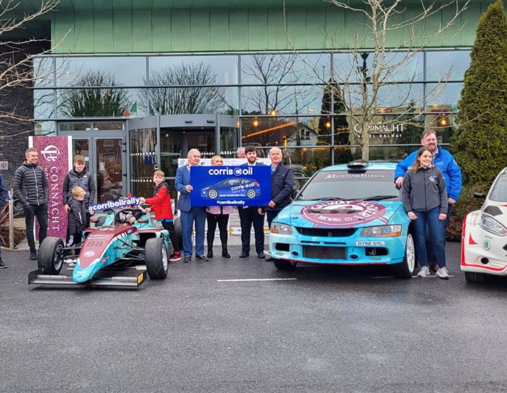 Galway International Rally Event Outside The Connacht Hotel – A group of people, including rally drivers, families, and event organizers, pose in front of The Connacht Hotel with two rally cars and a formula-style race car. The cars are painted in blue and purple with event branding. A banner for "Corrib Oil Rally" is held by officials, highlighting the hotel's involvement in the event.