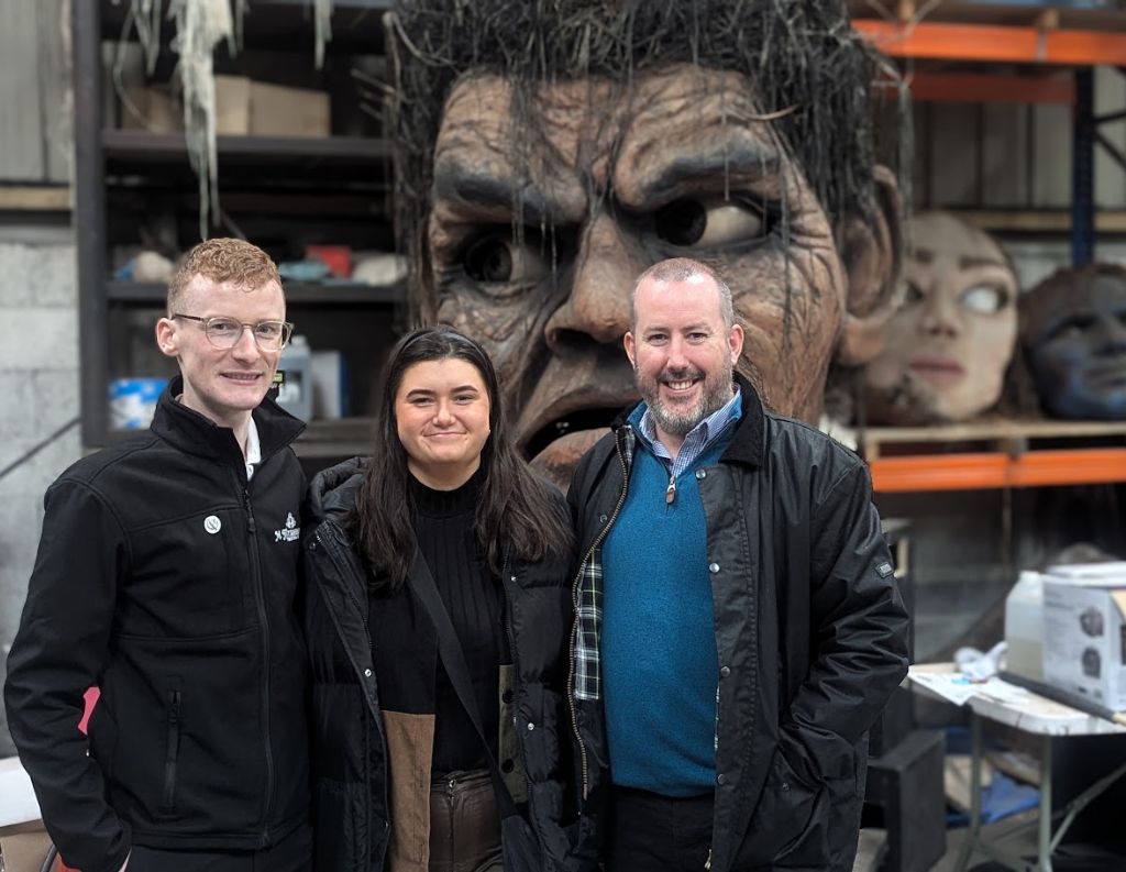 Three People Posing in a Workshop with Giant Heads in the Background – A group of three individuals stand smiling in front of a large, dramatic sculpture of a face, surrounded by other oversized artistic heads, inside an industrial-style workshop.