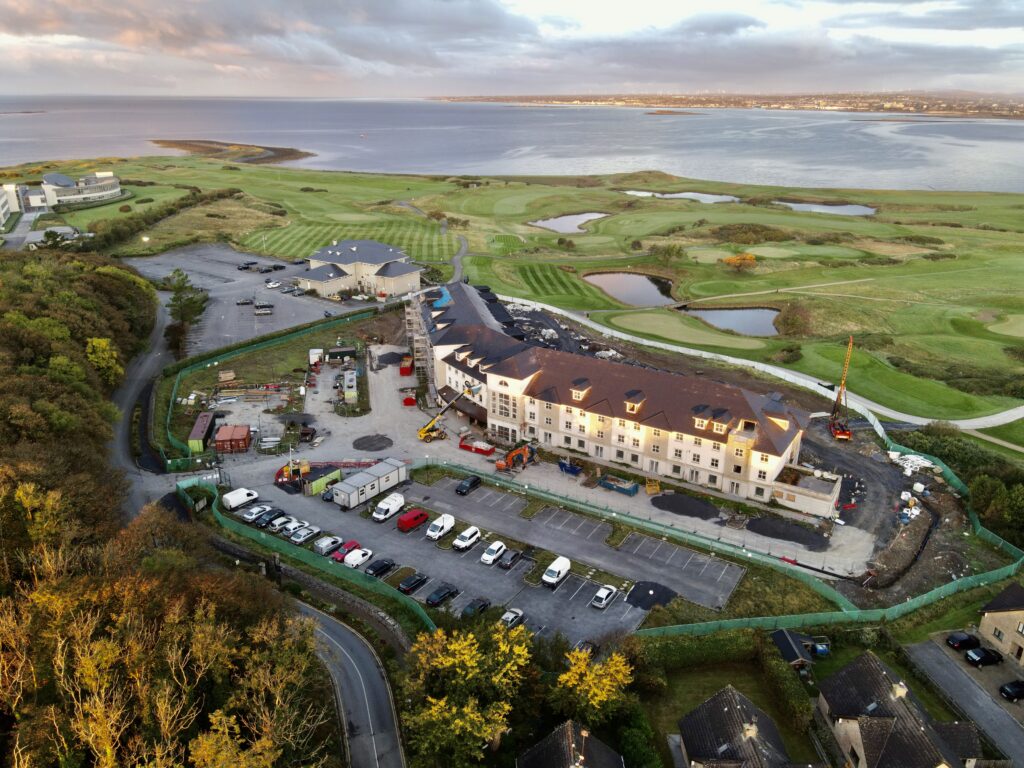 Coastal Hotel Development Progress – An aerial shot of a hotel under construction near a picturesque coastline, featuring a scenic golf course, ample parking, and stunning views of the water, with the golden hues of sunset illuminating the scene.