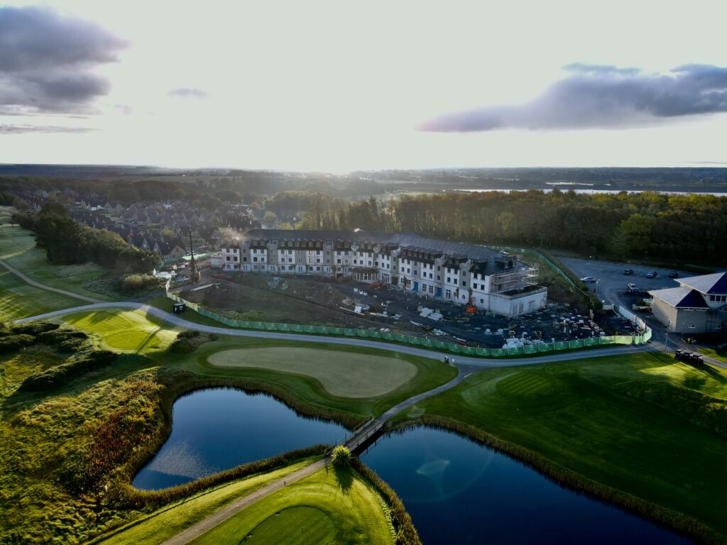 Aerial View of The Hawthorn Under Construction – A scenic aerial shot of a golf course with a partially built hotel or residential complex, surrounded by lush greenery and a reflective pond.