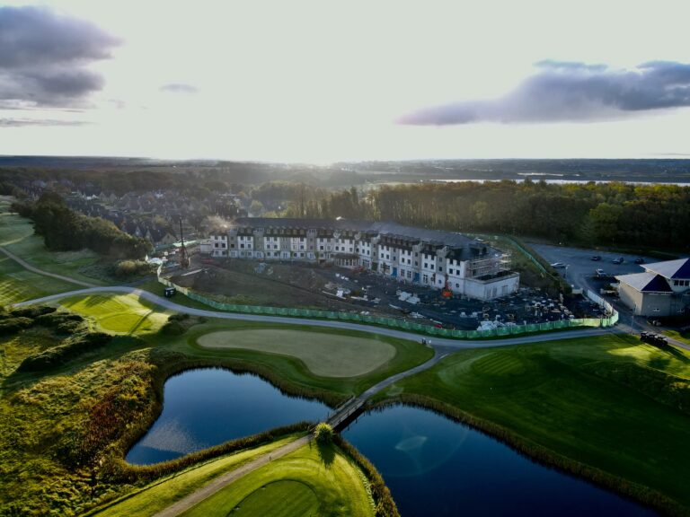 Aerial View of The Hawthorn Under Construction – A scenic aerial shot of a golf course with a partially built hotel or residential complex, surrounded by lush greenery and a reflective pond.