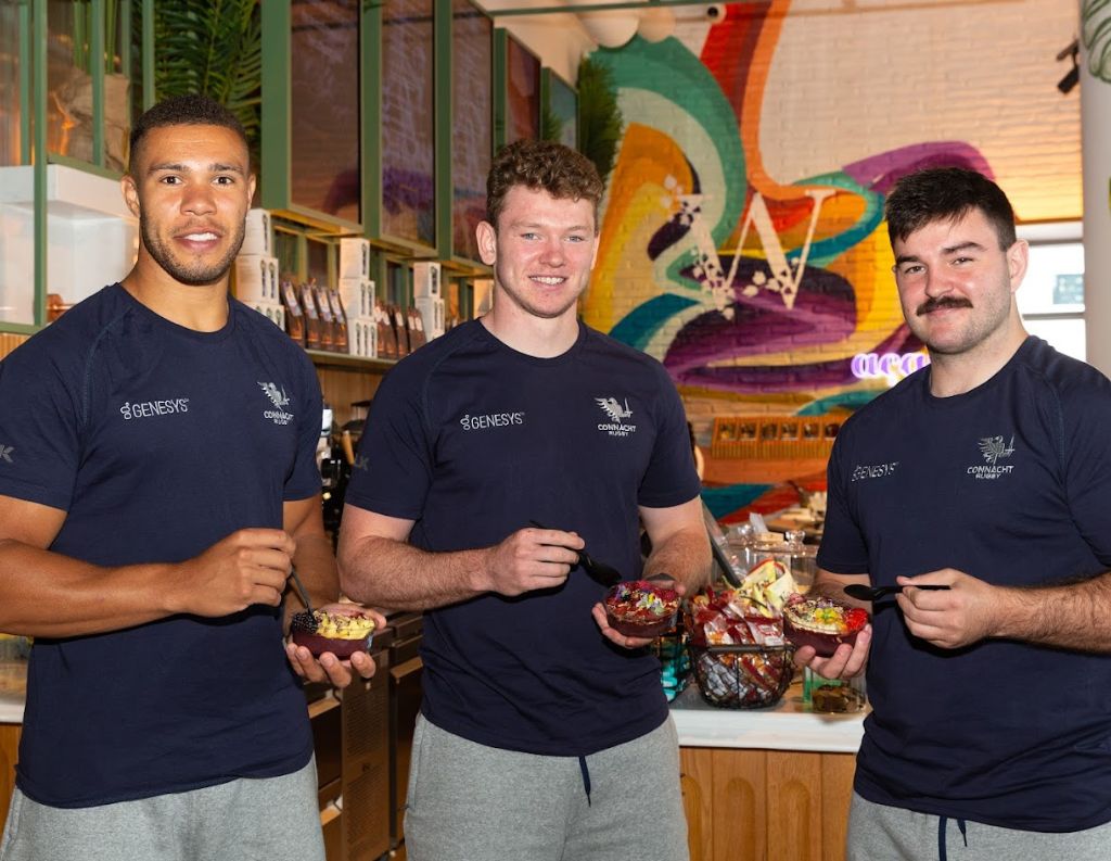 Connacht Rugby Players Enjoying a Healthy Meal – Three Connacht Rugby players, dressed in team shirts, smile while holding acai bowls inside a café with a colorful mural in the background, promoting a healthy lifestyle.