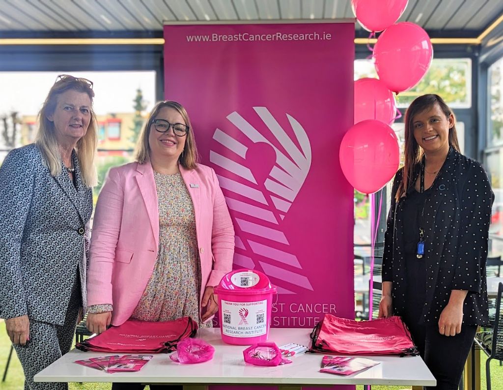 Breast Cancer Research Awareness Stand – Three women stand behind a bright pink Breast Cancer Research Institute promotional table, surrounded by pink balloons and fundraising materials.