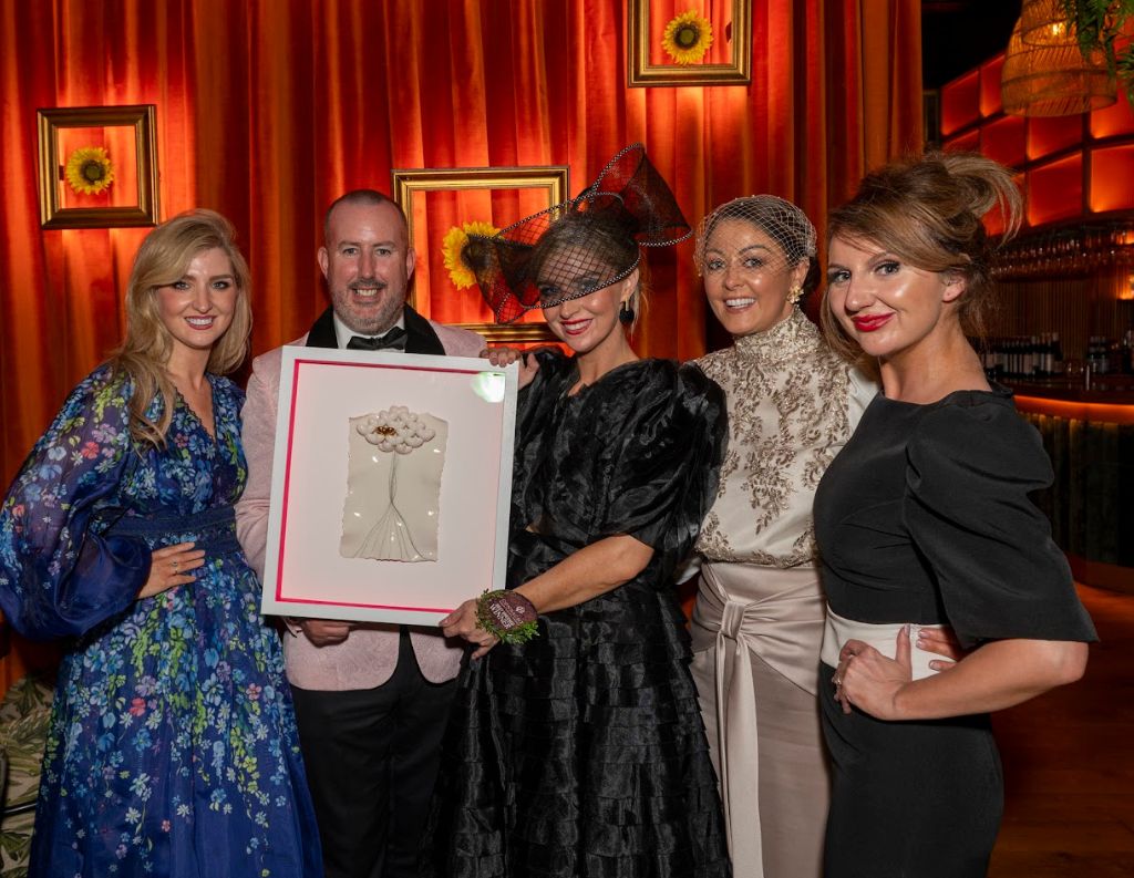 Elegant Group Holding an Award in a Stylish Venue – A group of five elegantly dressed individuals, including women in glamorous attire and fascinators, pose inside a chic venue with warm lighting and framed artwork. One person holds a framed award, showcasing a moment of celebration.