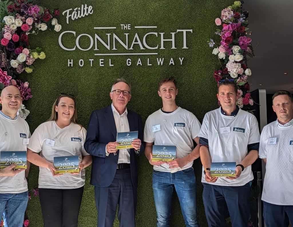 Connacht Hotel Team Gathering – A group of six people standing in front of a green floral backdrop at The Connacht Hotel Galway, holding booklets and dressed in branded t-shirts and business attire.