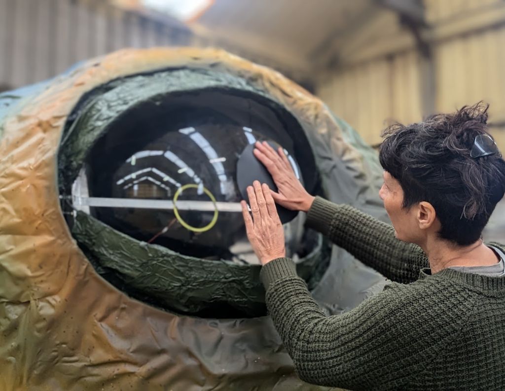 Artist Working on a Large Eye Structure – A person wearing a green sweater carefully adjusts a translucent panel on a giant, intricately designed eye structure, possibly part of an art installation or parade float, inside a workshop setting.