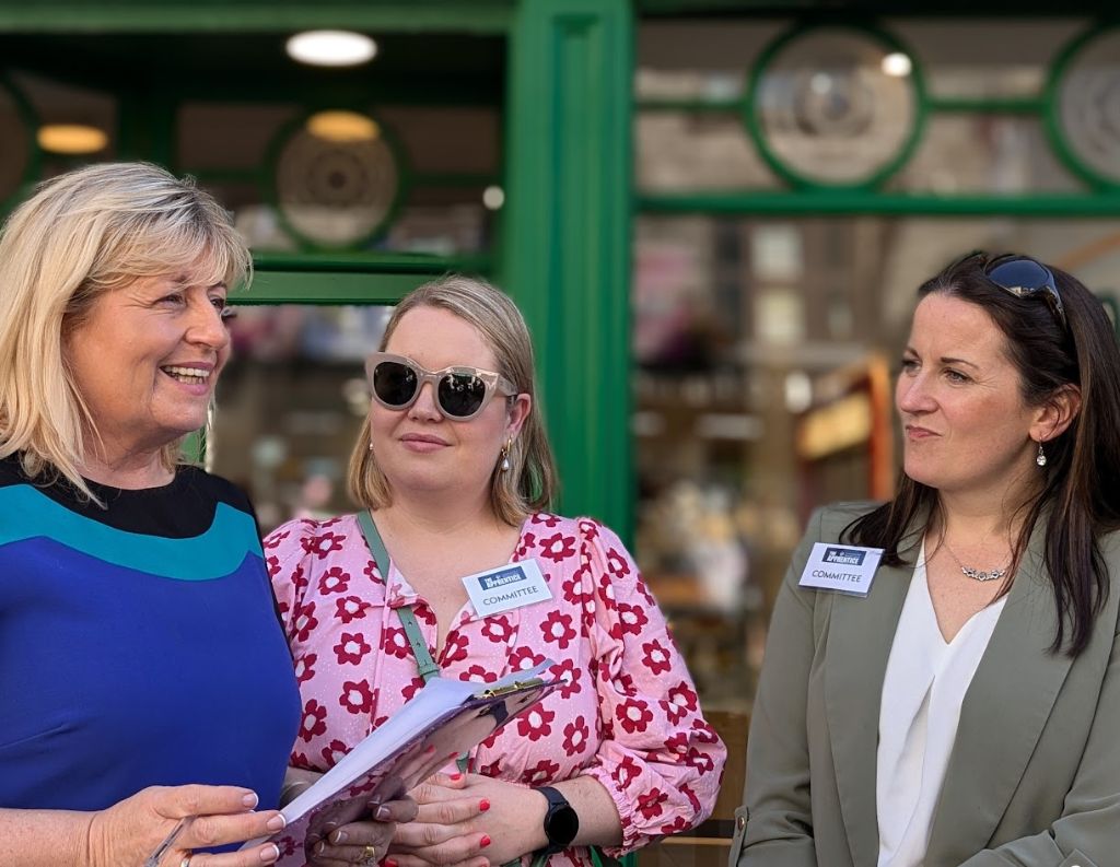 Women Networking at an Event – Three professionally dressed women in discussion at an outdoor event, one of them holding a clipboard, standing in front of a green storefront.