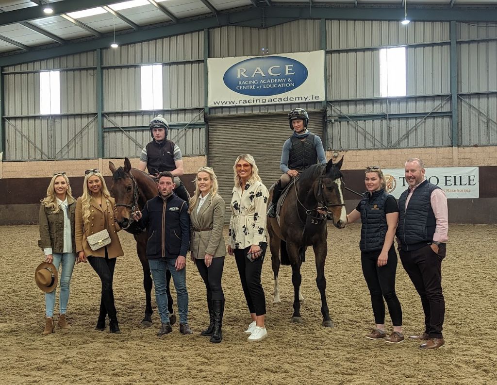 Equestrian Training Facility with Riders and Guests – A group of people, including two mounted riders and stylishly dressed guests, pose inside an indoor equestrian training facility with "RACE" branding, showcasing an event related to horse racing or training.