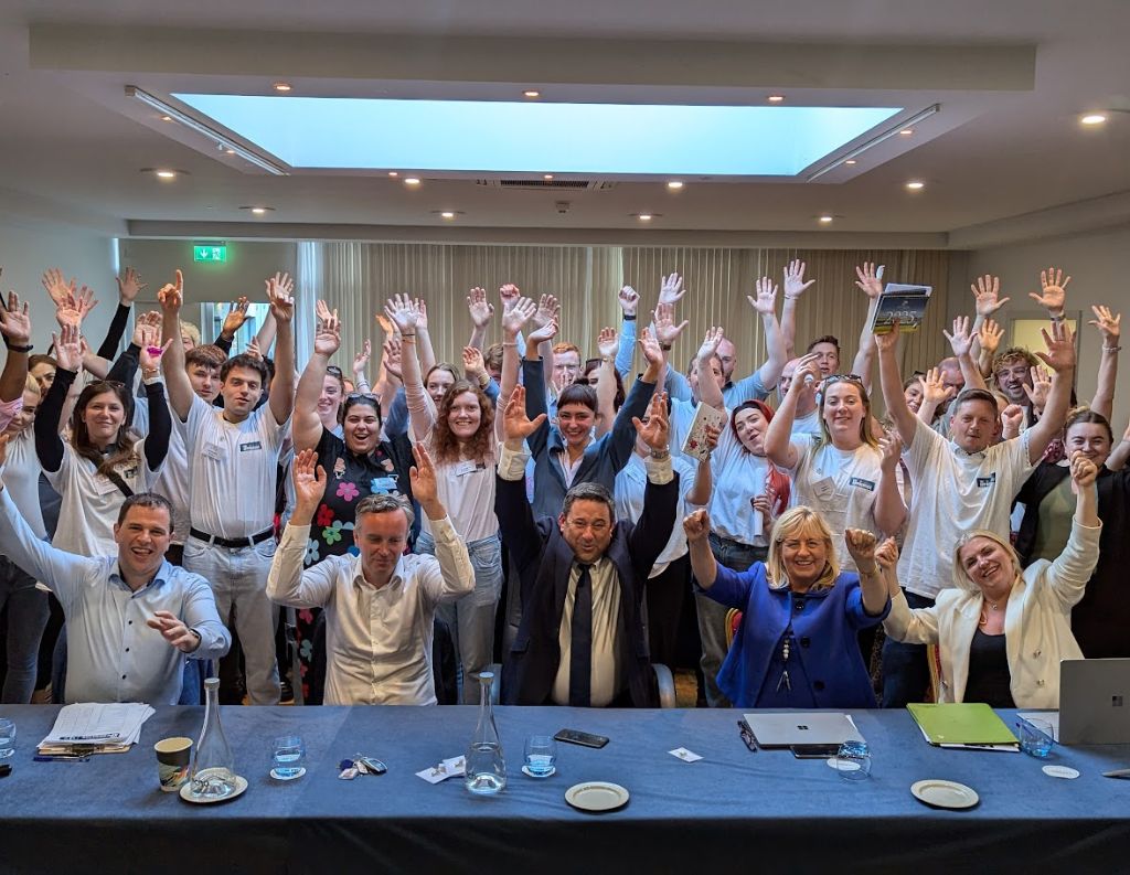 Energetic Group Conference – A large group of people in a conference room raising their hands enthusiastically, engaging in a business or hospitality event.