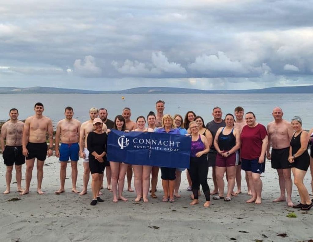 Connacht Hospitality Group Sea Swim Event – A large group of people standing together on the beach in swimwear, smiling and holding a Connacht Hospitality Group banner, celebrating a successful sea swim against the backdrop of the Atlantic Ocean and cloudy skies.