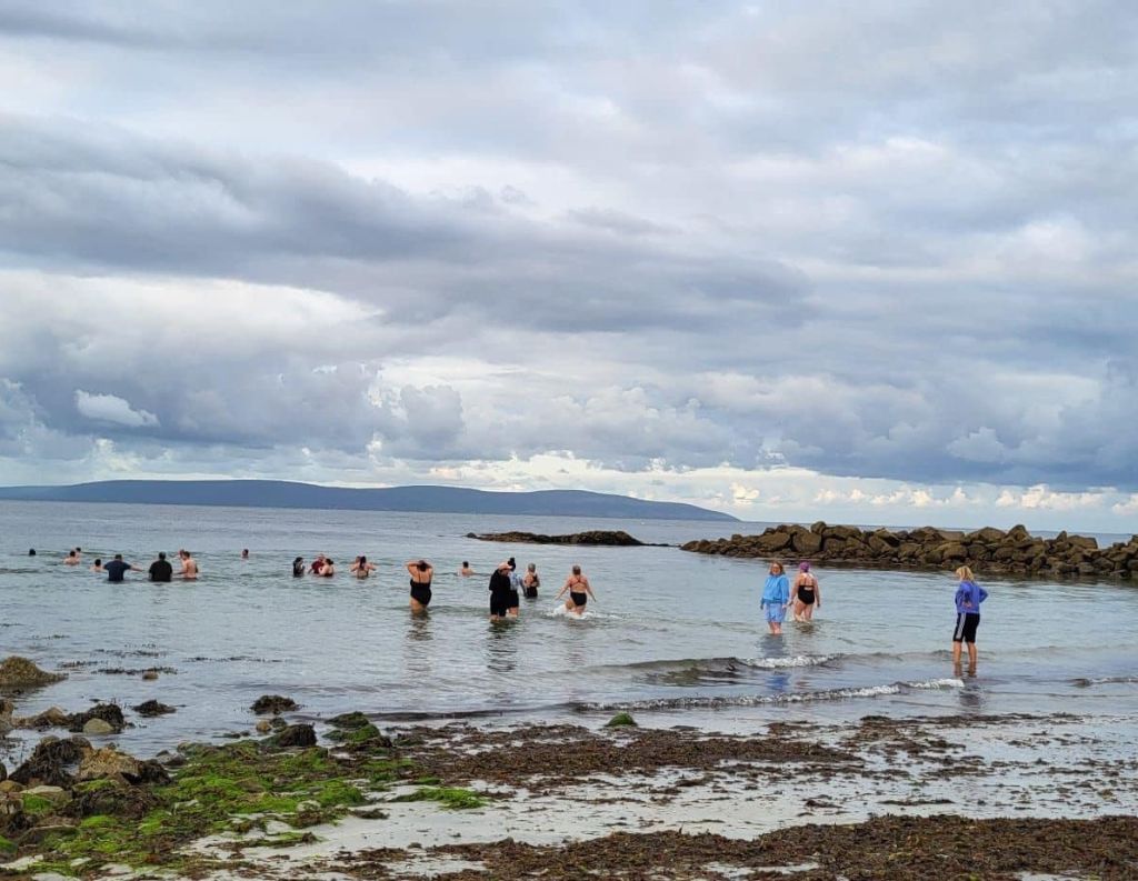 Group Sea Swimmers in Galway – A group of people wading into the sea at a Galway beach under a cloudy sky, participating in an open-water swim with a scenic backdrop of distant hills.