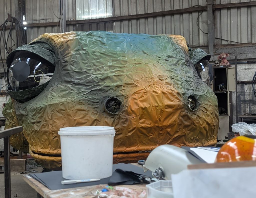 Massive Frog Sculpture Under Construction – A large, colorful frog-like structure with oversized eyes sits inside a workshop, surrounded by tools and materials, appearing to be part of a creative project or event display.