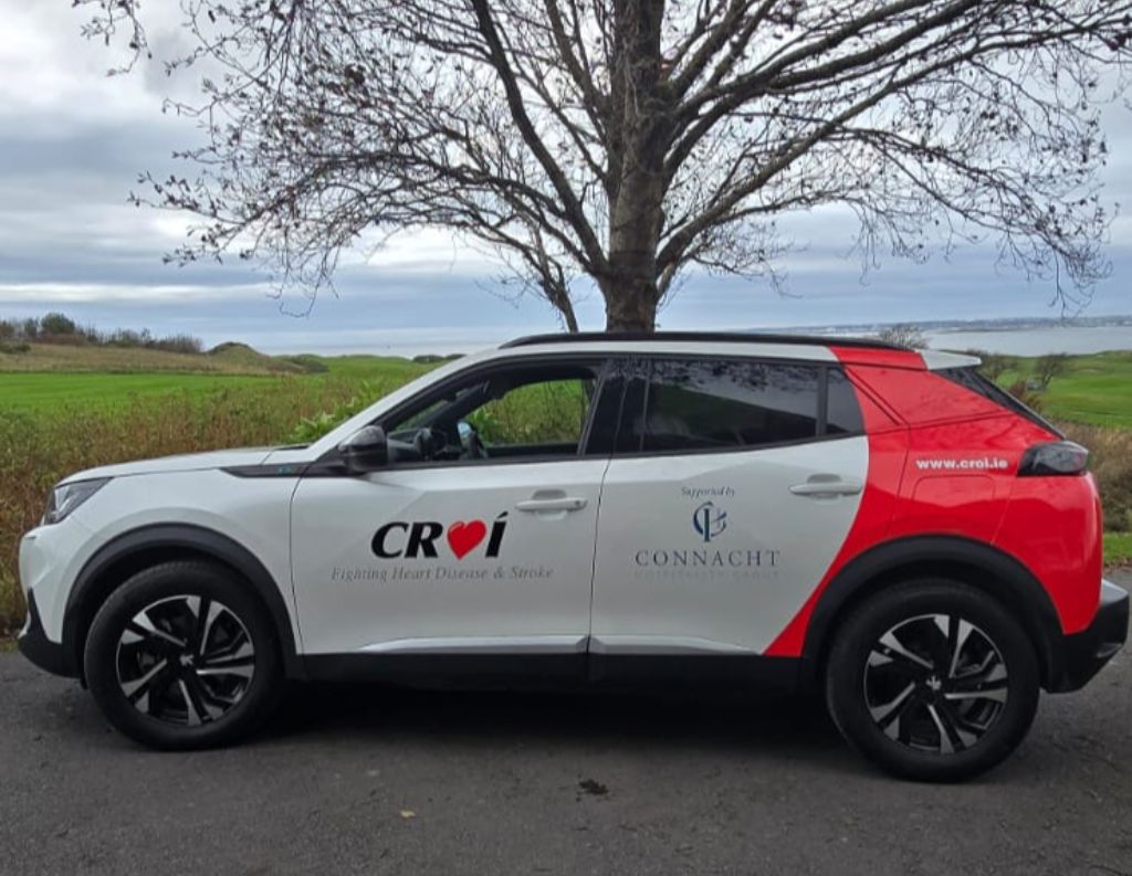 Croi Charity Car Sponsored by Connacht Hospitality Group – A branded white and red car with logos of Croí, a heart disease and stroke charity, and Connacht Hospitality Group, parked by a coastal landscape with green fields and a tree in the background.