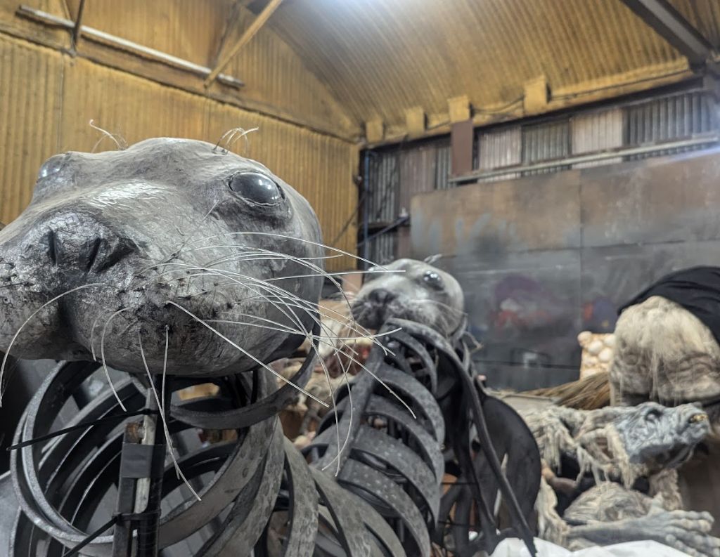 Mechanical Seal Sculptures in a Workshop – A hauntingly detailed pair of seal sculptures made from metal and synthetic materials, with exposed mechanical elements, rest inside a dimly lit workshop, likely part of a larger artistic or theatrical installation.