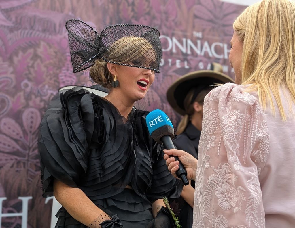 Woman in Elegant Black Outfit Being Interviewed by RTÉ – A woman in a striking black dress with a dramatic fascinator speaks into an RTÉ microphone while being interviewed at a Connacht Hotel-branded backdrop, highlighting a high-profile fashion or racing event.