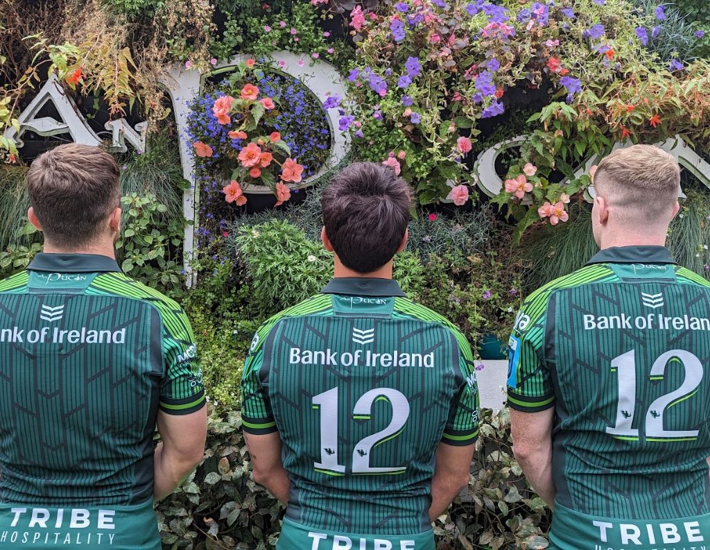 Connacht Rugby Players Facing a Floral Wall at An Púcán – Three Connacht Rugby players stand with their backs to the camera, wearing matching green jerseys with the number 12, in front of a flower-covered An Púcán sign, showcasing their team unity and support.