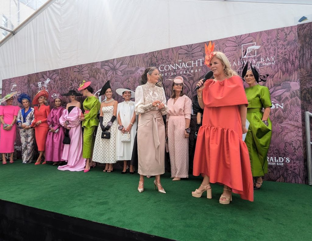 Best Dressed Event at Galway Races – A lineup of stylishly dressed women in bold colors and elegant fascinators stand on a stage with a Connacht Hotel-branded backdrop, while two hosts, also in fashionable attire, engage the crowd.
