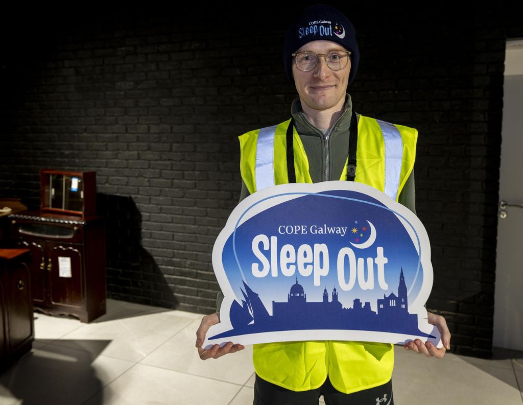 COPE Galway Sleep Out Advocate – A man in a high-visibility vest and beanie holds a COPE Galway "Sleep Out" sign, showing his commitment to the charity’s cause in a warmly lit indoor setting.