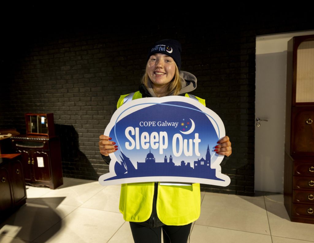 COPE Galway Sleep Out Participant – A smiling woman wearing a high-visibility vest and beanie holds a COPE Galway "Sleep Out" sign, promoting the charity’s initiative to support those experiencing homelessness.