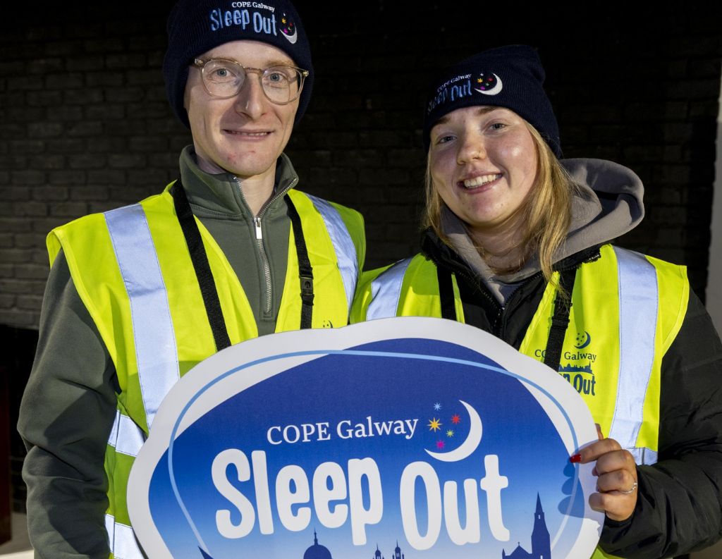 Two volunteers wearing high-visibility vests and COPE Galway beanies smile while holding a "COPE Galway Sleep Out" sign, supporting the charity's initiative to raise awareness for homelessness.