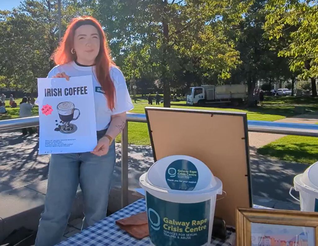 Fundraising for Galway Rape Crisis Centre – A young woman with red hair, wearing a white branded t-shirt, holds an "Irish Coffee" sign at an outdoor fundraising stall, which includes donation buckets labeled for the Galway Rape Crisis Centre.