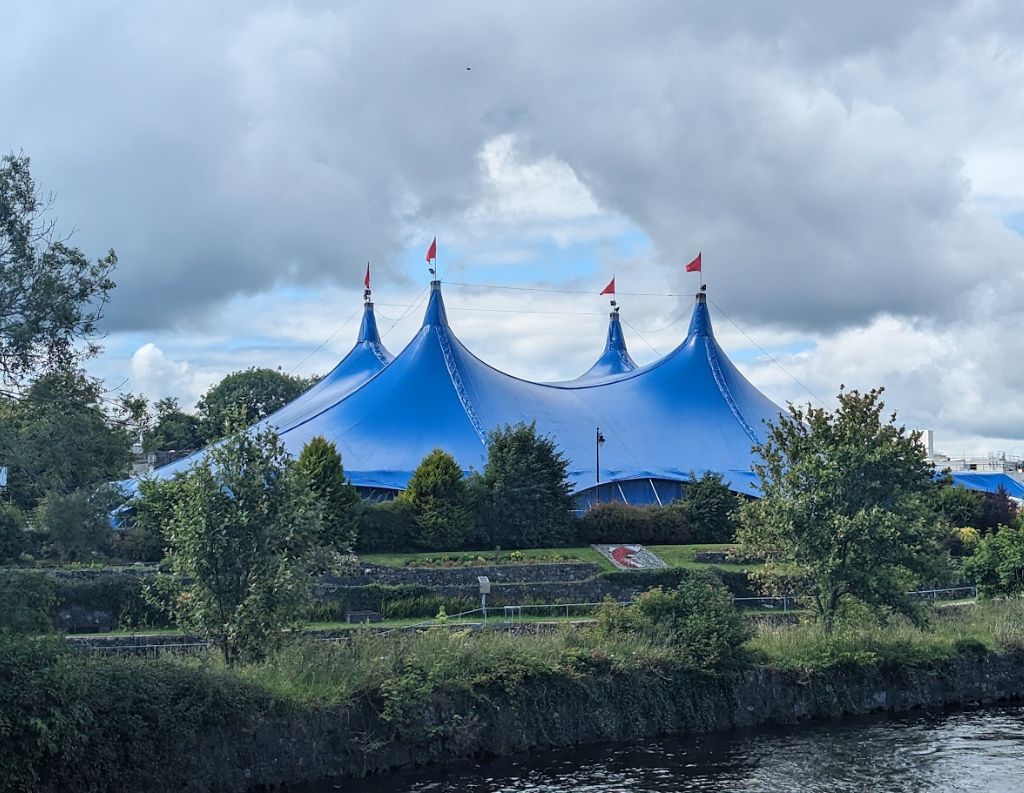 Large Blue Festival Tent at Galway International Arts Festival – A striking blue tent with multiple peaks and red flags stands in a green park-like setting, marking a major venue for the Galway International Arts Festival.