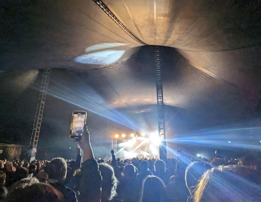Concert Inside the Big Top Tent at Galway International Arts Festival – A crowd inside the large festival tent enjoys a concert, with stage lights beaming across the darkened space. A person holds up a phone to capture the performance.