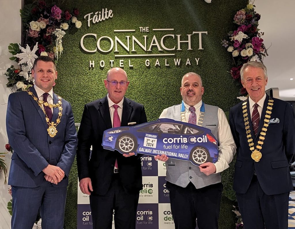 Galway International Rally Launch Event at The Connacht Hotel – Four distinguished men, including local officials wearing ceremonial chains, pose in front of a green floral wall with "The Connacht Hotel Galway" signage. One man holds a cutout of a rally car with "Corrib Oil Galway International Rally" branding, celebrating the launch of the prestigious motorsport event.