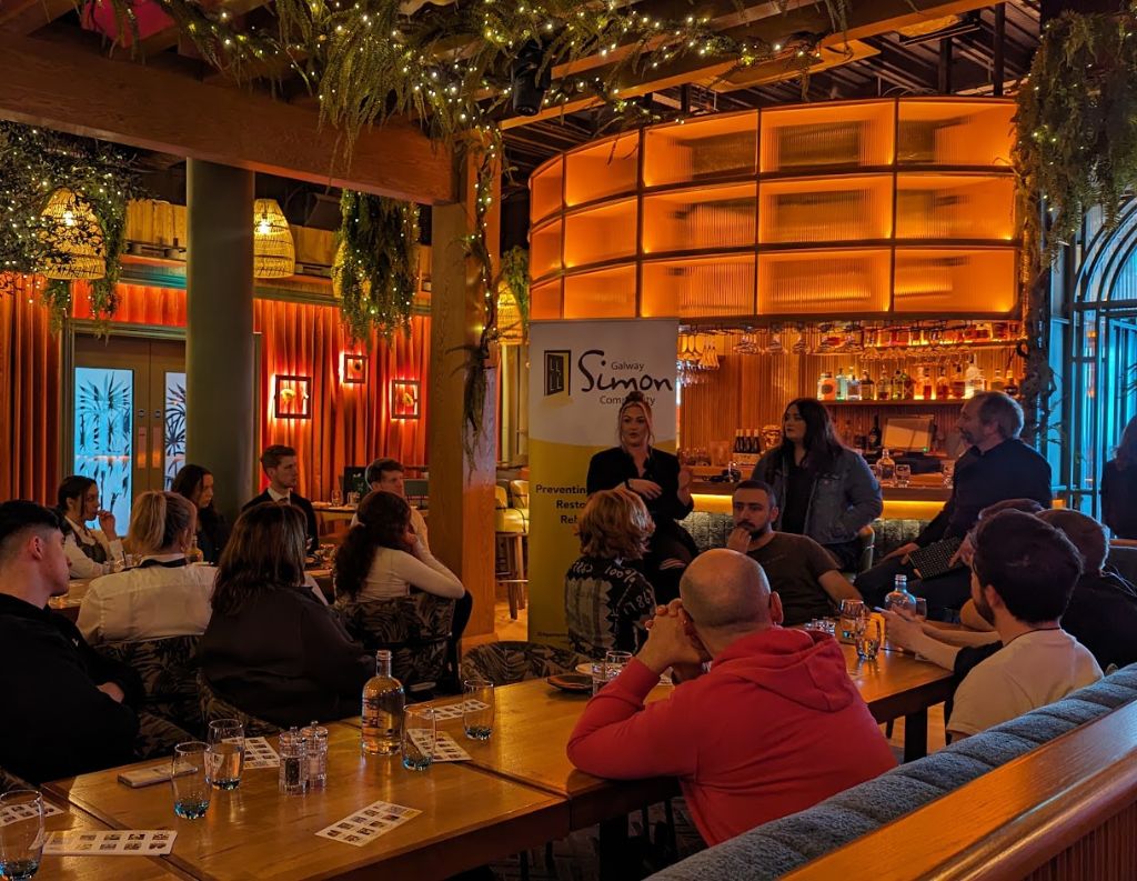 Galway Simon Community Awareness Talk – A panel discussion takes place in a cozy venue, where speakers discuss homelessness prevention and support, with attendees listening intently. A Galway Simon Community banner is visible in the background.