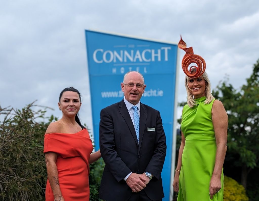 Three well-dressed individuals, including a man in a suit and two women in vibrant dresses with stylish fascinators, pose in front of a "Connacht Hotel" banner, highlighting a formal event setting.