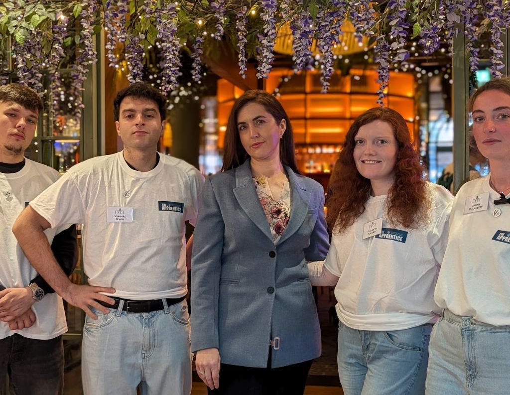 Empowered Team at a Community Event – A confident woman in a blazer stands alongside four young volunteers wearing white t-shirts with name badges, posing in an elegant venue decorated with hanging flowers.