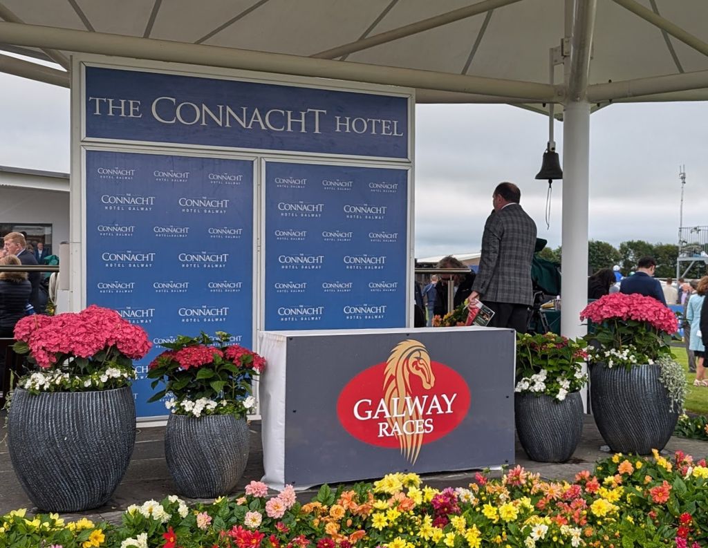 Connacht Hotel Branded Stage at Galway Races – A stage featuring "The Connacht Hotel" branding and a "Galway Races" sign, surrounded by vibrant flowers. A man stands near the podium, emphasizing the connection between the hotel and the prestigious horse racing event.